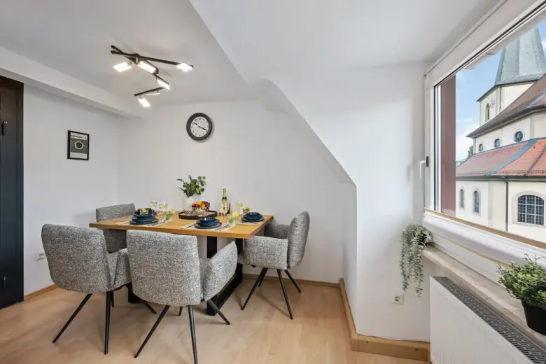 Der moderne Essbereich in App.3 besteht aus einem Holztisch für sechs Personen, grauen gepolsterten Stühlen und Dekoration darüber. Das Fenster mit Blick auf die Bachstraße zeigt eine Kirche und eine Topfpflanze steht auf der Fensterbank.