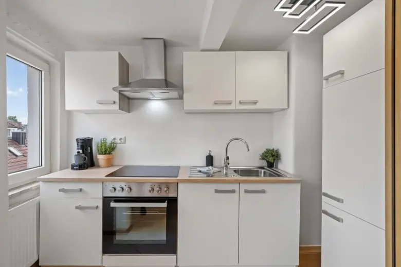 Moderne, kompakte Küche in der Bachstraße (App.3.) mit weißen Schränken, Elektroherd, Backofen, Dunstabzugshaube aus Edelstahl, Spüle, Kaffeemaschine, Topfpflanzen und einem Fenster, das Tageslicht hereinlässt. Minimalistisches und klares Design.