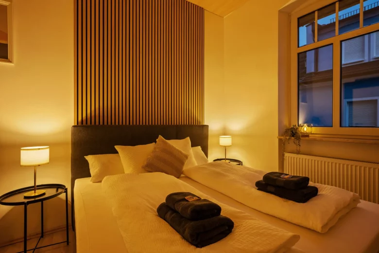 Ein gemütliches, warm beleuchtetes Schlafzimmer mit einem Doppelbett, zwei Nachttischen mit Lampen, gefalteten Handtüchern auf dem Bett, Wanddekoration aus Holzlatten hinter dem Kopfteil und einem großen Fenster mit Blick auf die Gebäude draußen.