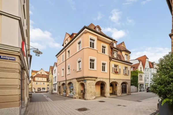 Ein pfirsichfarbenes Gebäude mit gewölbten Steintüren steht an einer ruhigen Straßenecke in einer europäischen Stadt und bietet charmante Ferienwohnungen Bad Kissingen inmitten von kopfsteingepflasterten Wegen, pastellfarbenen Gebäuden und einem klaren blauen Himmel.