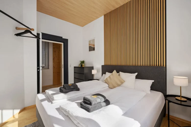 Modernes Schlafzimmer in einem Bad Kissinger Appartement mit großem Bett, ordentlich gefalteten Handtüchern, Holzlattenwand, schwarzer Kommode und minimalistischer Einrichtung. Das natürliche Licht aus dem Fenster unterstreicht die gemütliche und saubere Atmosphäre in der Bachstraße.