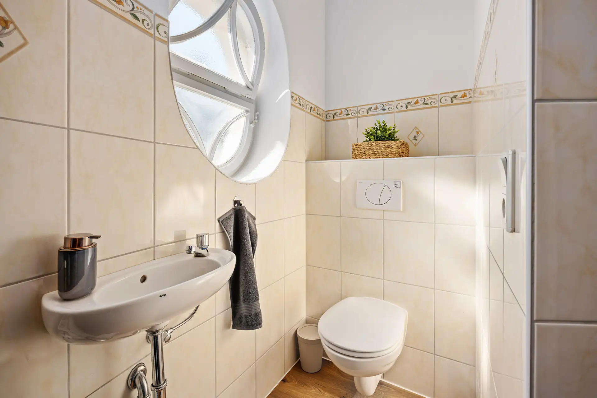 Ein kleines, gefliestes Bad in einer Bad Kissinger Ferienwohnung mit einem Waschbecken an der Wand, einem grauen Handtuch daneben, einer modernen Toilette, einer kleinen Pflanze auf einem Regal über der Toilette und einem runden Fenster, das Tageslicht hereinlässt.