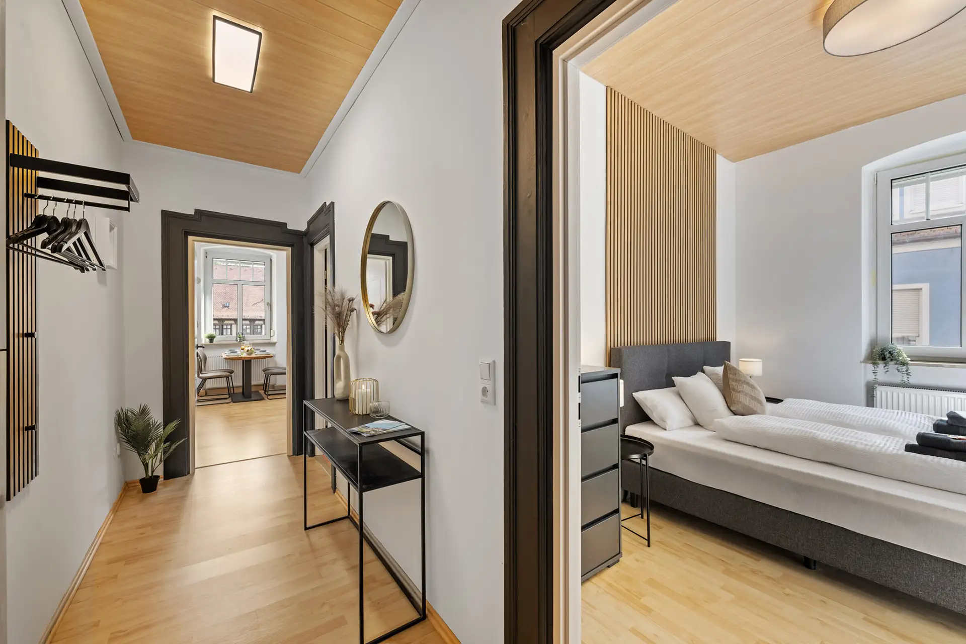 Eine helle, moderne Ferienwohnung in der Bachstraße mit hellen Holzböden und einer hellen Decke, die zu einem Schlafzimmer mit einem großen Bett und einer neutralen Einrichtung führt, sowie einem Essbereich, der im Hintergrund sichtbar ist.