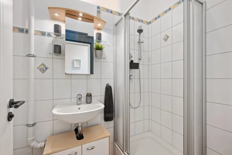 Ein modernes Badezimmer in einer Bad Kissinger Ferienwohnung verfügt über weiß geflieste Wände, eine Eckdusche mit Glastüren, ein kompaktes Waschbecken mit Unterschrank, einen Spiegel mit Ablageflächen und stilvolle Dekorationsartikel wie eine Pflanze und Handtücher.