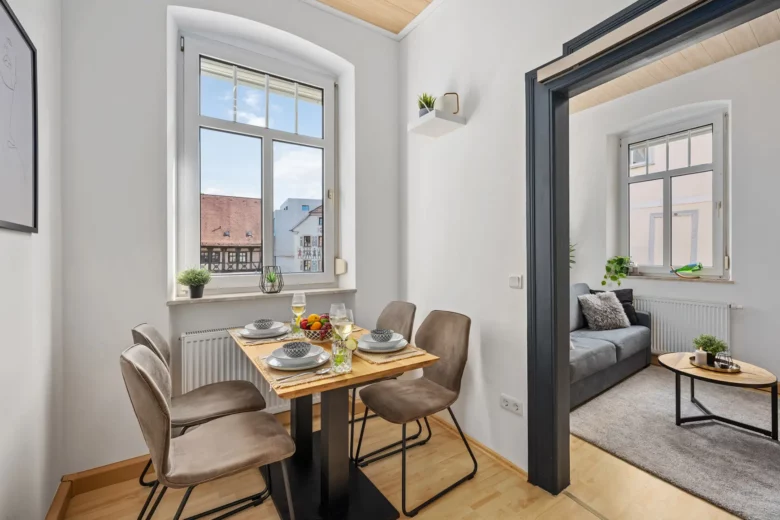 Ein moderner Appartment-Essbereich mit einem Holztisch für vier Personen, braunen Stühlen und einem Fenster mit Blick auf die Bachstraße. Im Hintergrund befindet sich ein gemütliches Wohnzimmer mit einem grauen Sofa und natürlichem Lichteinfall.