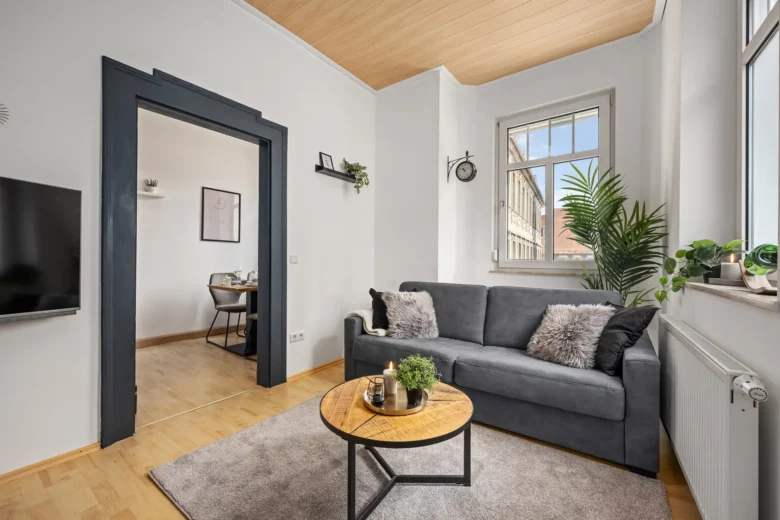 Modernes Ferienwohnung-Wohnzimmer in der Bachstraße in Bad Kissingen mit grauem Sofa, flauschigen Kissen, rundem Holzcouchtisch, Topfpflanzen, Wandkunst und großen Fenstern. Helle Holzböden und ein Essbereich vervollständigen den hellen, einladenden Raum.