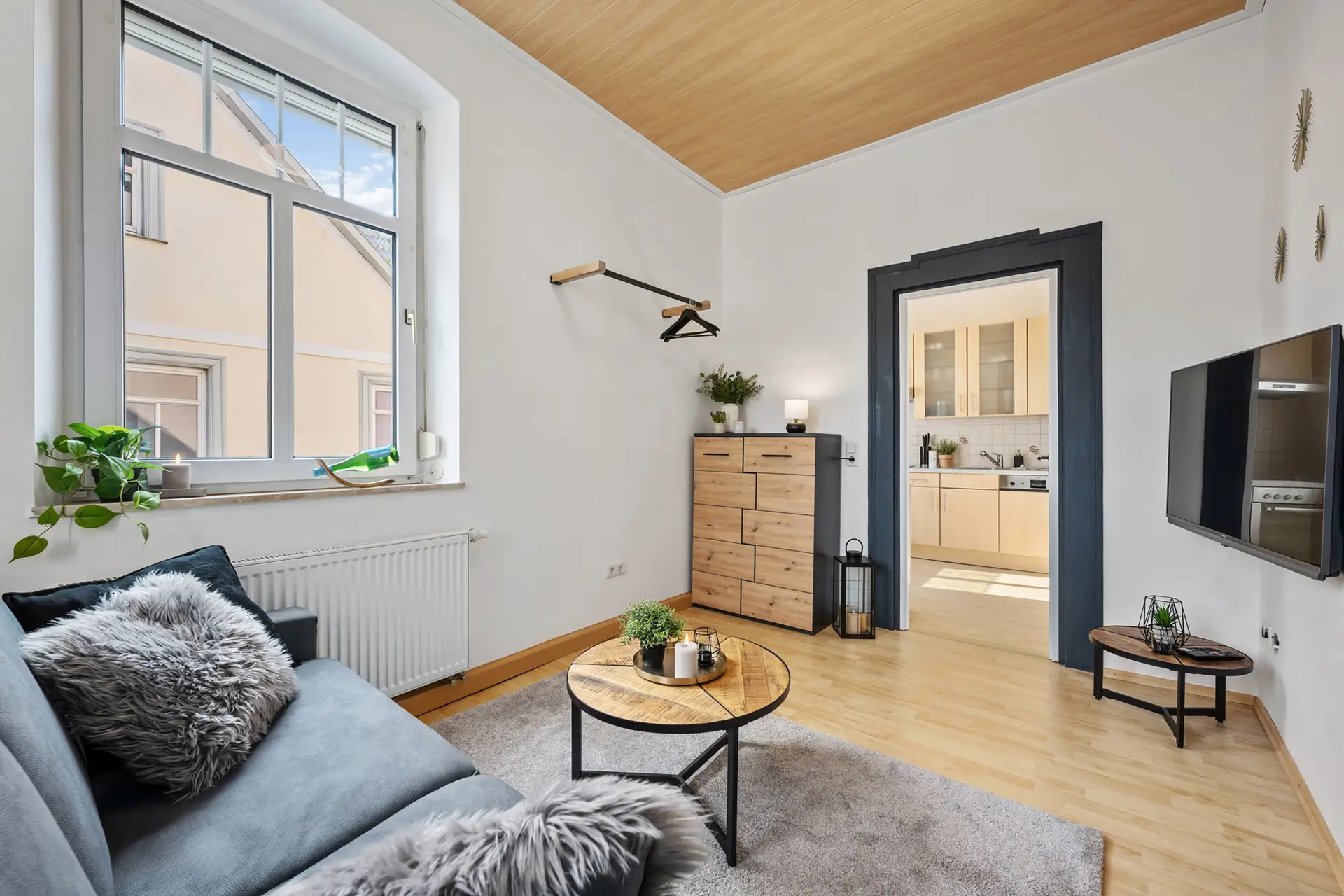 Helles, modernes Wohnzimmer in der Ferienwohnung in Bad Kissingen mit hellem Holzboden, grauem Sofa mit Fellkissen, rundem Couchtisch, Kommode, Wandfernseher, Pflanzen und offenem Durchgang zur Küche mit hellen Schränken in der Bachstraße.