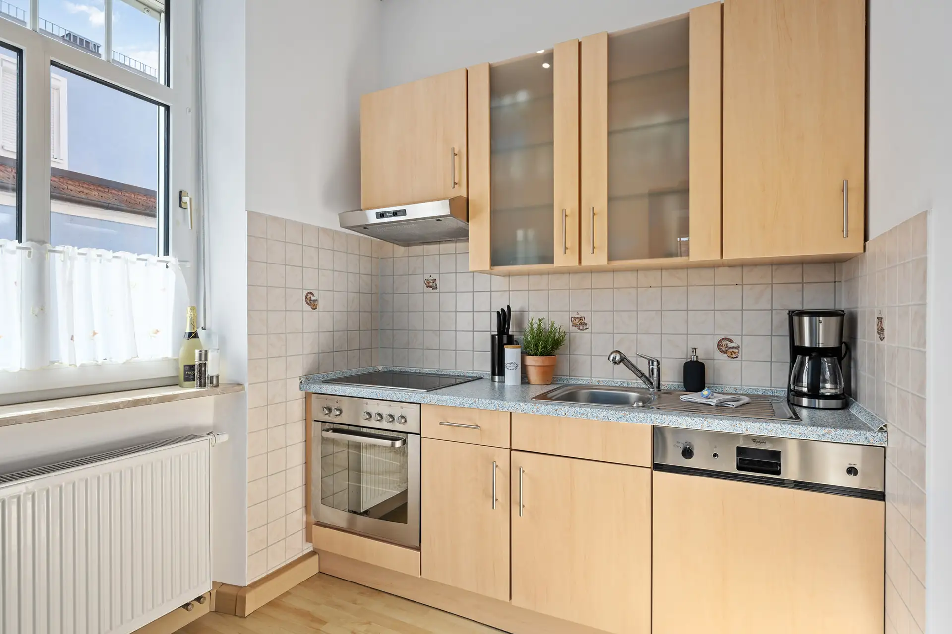 Eine kleine, moderne Ferienwohnung-Küche mit hellen Holzschränken, Herd, Backofen, Spüle, Geschirrspüler, Kaffeemaschine und Topfpflanze. Sonnenlicht strömt durch ein großes Fenster mit einem weißen Vorhang auf die hellen Fliesen und den Boden.