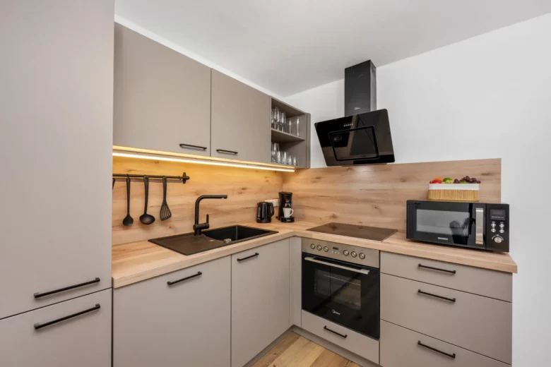 Moderne Küche mit hellgrauen Schränken, schwarzer Spüle, eingebautem Ofen, Herd, Mikrowelle, Kaffeemaschine und an der Wand hängenden Utensilien. Arbeitsplatte und Aufkantung aus Holz, mit Schale mit Obst auf der Theke.