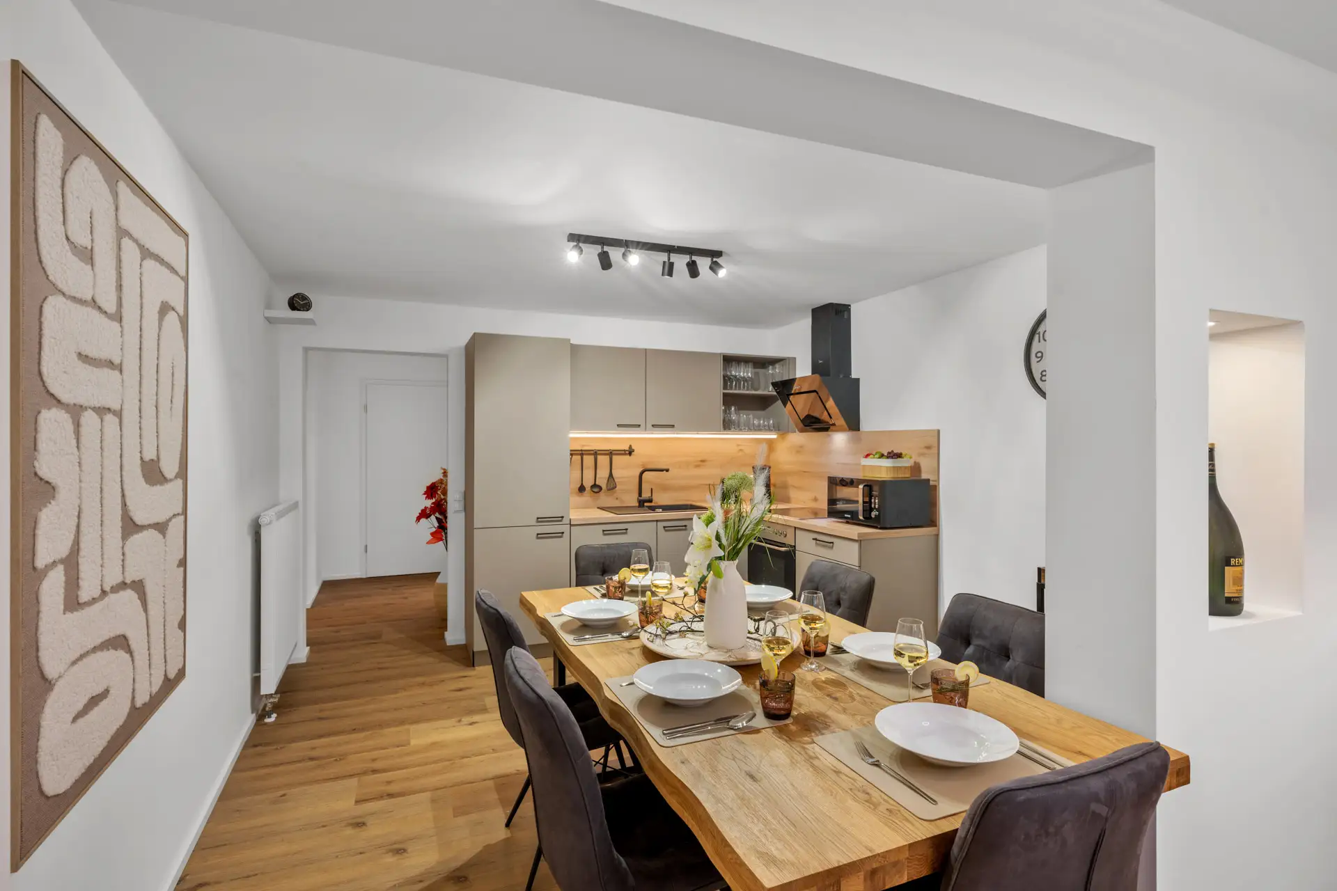 Modernes Esszimmer und Küche mit Holzböden, einem großen Holztisch für sechs Personen, abstrakter Wandkunst, schlichten Schränken, offenen Regalen und sanfter Beleuchtung, die eine gemütliche, einladende Atmosphäre schaffen.