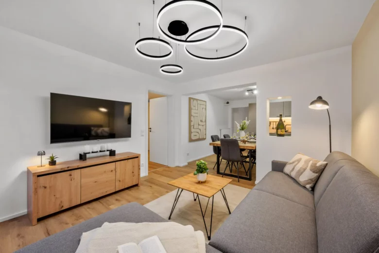 Modernes Wohnzimmer mit einem grauen Sektionssofa, einem Couchtisch aus Holz, einem Flachbildfernseher auf einem Holzschrank, runden Deckenleuchten und einem offenen Essbereich im Hintergrund mit einem gedeckten Tisch für eine Mahlzeit.