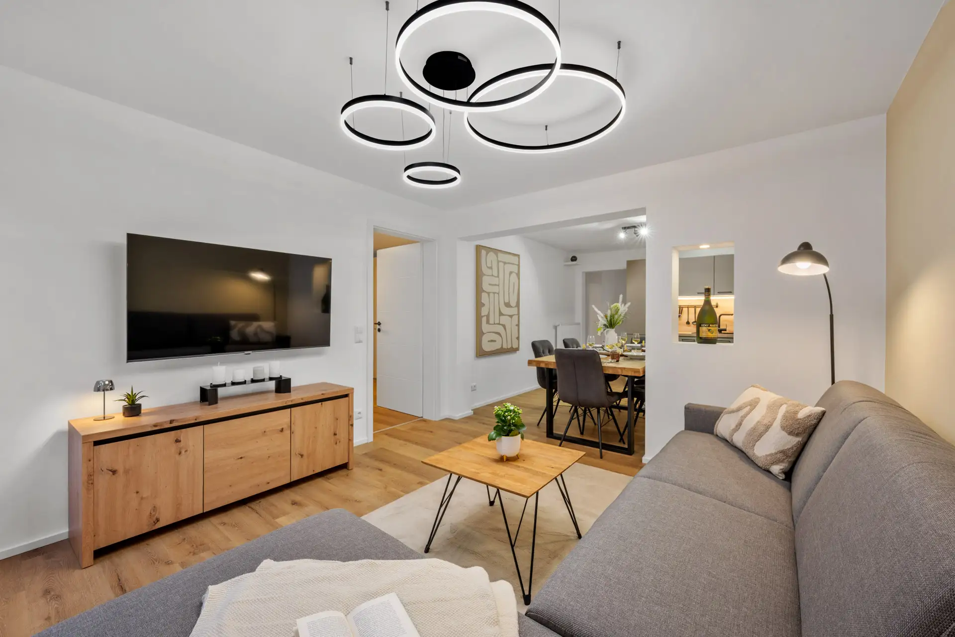 Modernes Wohnzimmer mit einem grauen Sektionssofa, einem Couchtisch aus Holz, einem Flachbildfernseher auf einem Holzschrank, runden Deckenleuchten und einem offenen Essbereich im Hintergrund mit einem gedeckten Tisch für eine Mahlzeit.