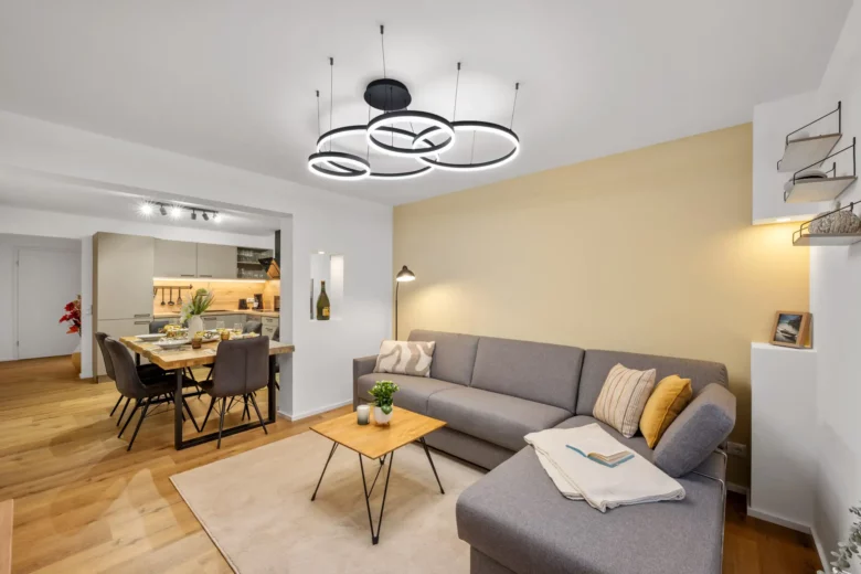 Modernes Wohnzimmer mit einem grauen Sektionssofa, einem Couchtisch aus hellem Holz und einer beigen Akzentwand, angrenzend an einen Essbereich mit gedecktem Tisch und offener Küche. Stilvolle runde Deckenleuchten erhellen den Raum.