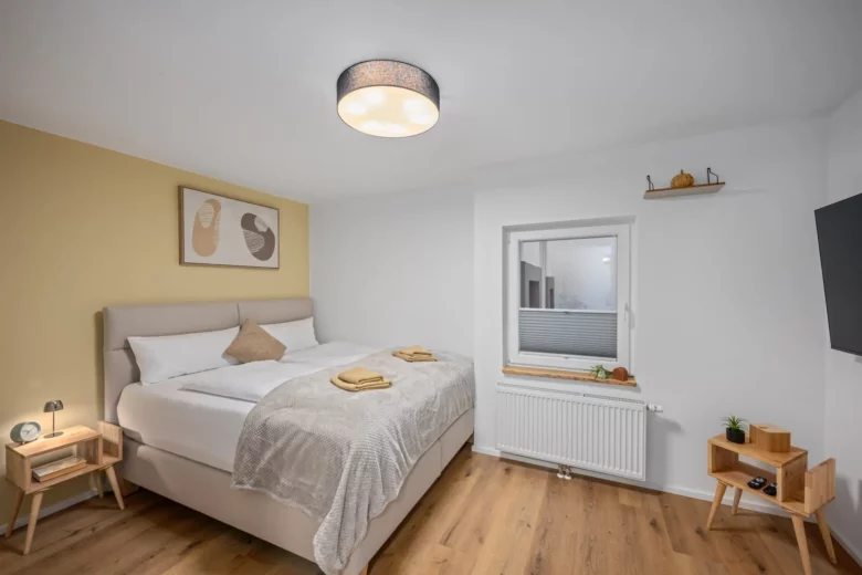 Ein modernes Schlafzimmer mit einem Doppelbett, heller Bettwäsche, gelber Akzentwand, Nachttischen mit Lampen, Holzboden, Kunstwerken über dem Bett, einem Fenster mit einem Heizkörper darunter, Regalen mit Dekoration und einem an der Wand montierten Fernseher.