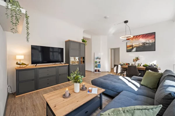 Modernes Appartment in Bad Kissingen mit großem grauen Sektionssofa, Couchtisch aus Holz, TV-Konsole, grünem Dekor und Essecke mit Stadtbildfoto. Sonnenlicht erfüllt den hellen Wohnbereich der Ferienwohnung.
