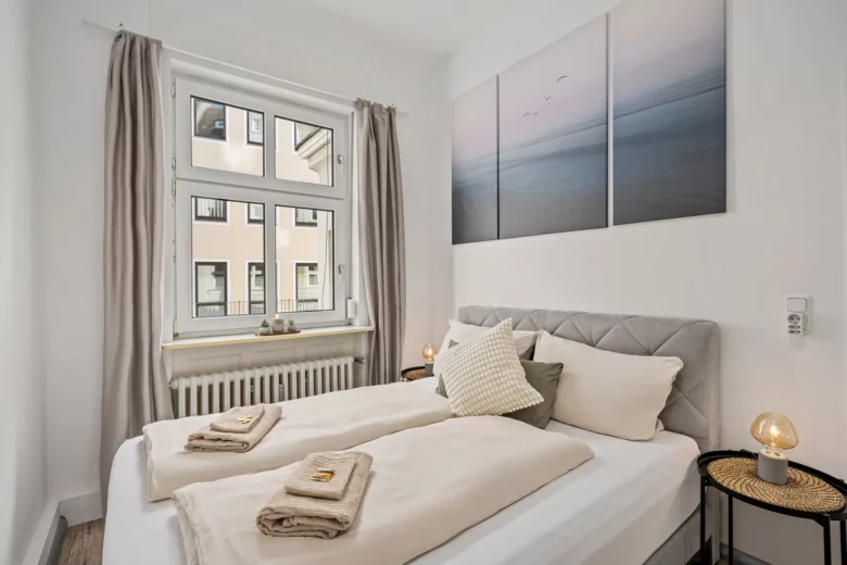 Eine gemütliche, moderne Ferienwohnung in der Unteren Marktstraße mit einem sauber gemachten Doppelbett mit beiger Bettwäsche. Handtücher liegen gefaltet auf dem Bett, es gibt ein großes Fenster, Wandkunst über dem Bett und zwei kleine Nachttische mit Lampen.
