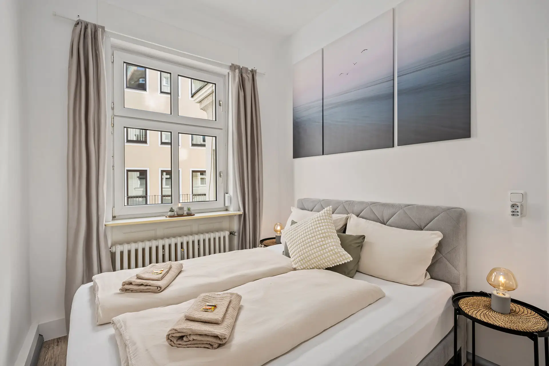 Eine gemütliche, moderne Ferienwohnung in der Unteren Marktstraße mit einem sauber gemachten Doppelbett mit beiger Bettwäsche. Handtücher liegen gefaltet auf dem Bett, es gibt ein großes Fenster, Wandkunst über dem Bett und zwei kleine Nachttische mit Lampen.