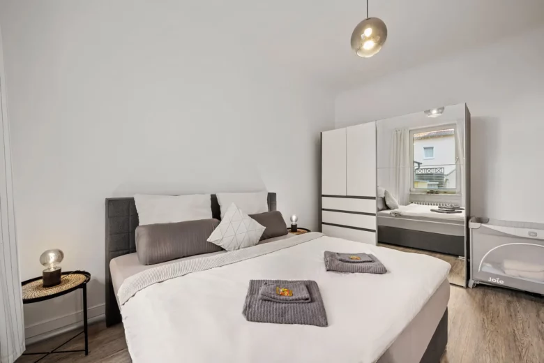 Ein modernes Schlafzimmer der Ferienwohnung Bad Kissingen mit einem Doppelbett, grauer und weißer Bettwäsche, gefalteten Handtüchern, Nachttischen mit Lampen, einem verspiegelten weißen Kleiderschrank und einem Kinderbett am Fenster. Der helle Raum ist mit Holzfußboden ausgestattet.