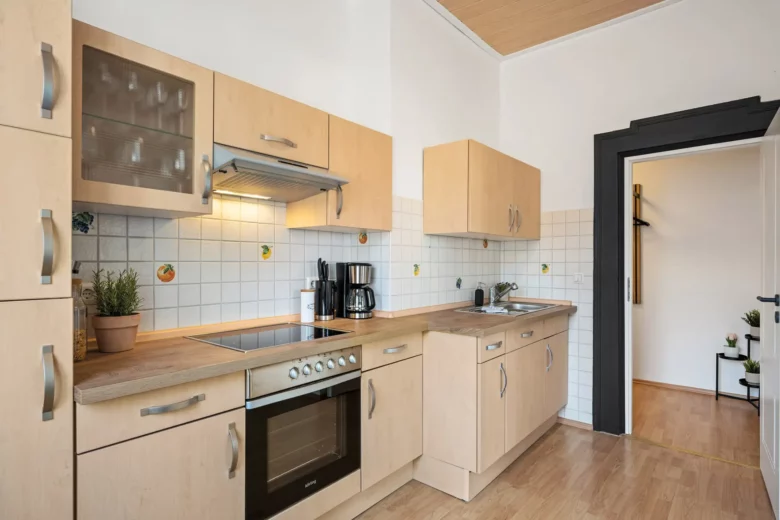 Moderne Küche in einer Ferienwohnung in Bad Kissingen mit hellen Holzschränken, Elektroherd, Backofen, Kaffeemaschine, Topfpflanze, Edelstahlspüle, gefliester Arbeitsplatte, Holzboden, weißen Wänden und einer schwarz gerahmten Tür.