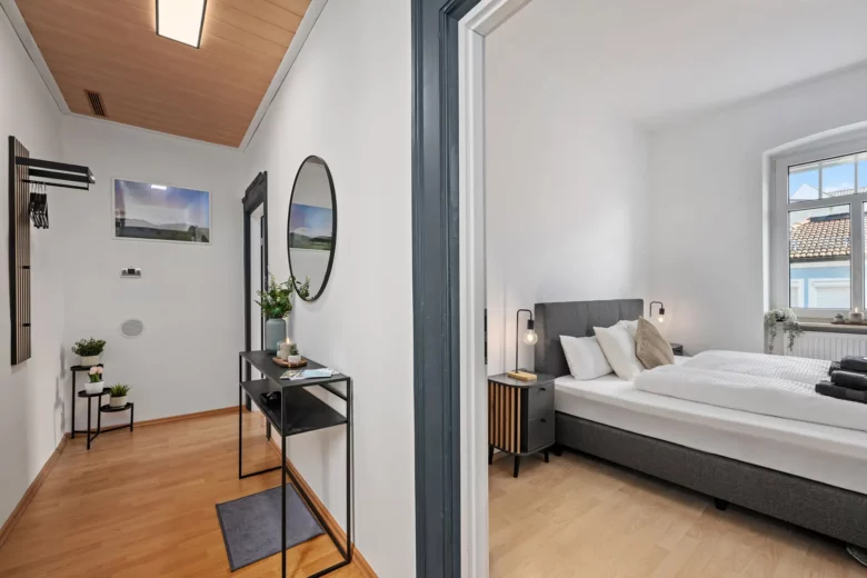 Die moderne Ferienwohnung in der Bachstraße verfügt über eine Diele mit Konsolentisch und Spiegel sowie ein angrenzendes Schlafzimmer mit Doppelbett, Nachttischen, Lampen und einem großen Fenster, das viel Tageslicht hereinlässt.