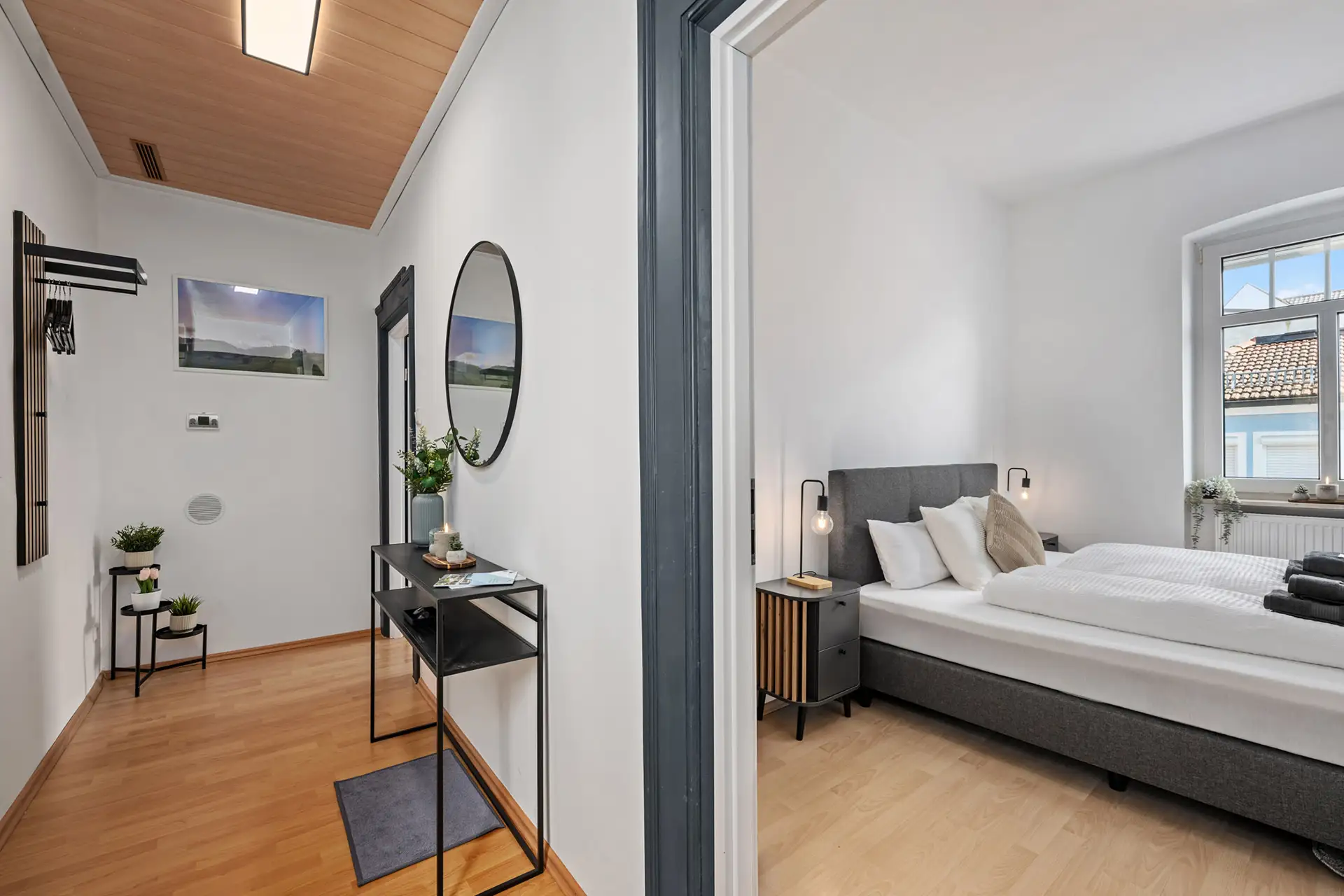 Die moderne Ferienwohnung in der Bachstraße verfügt über eine Diele mit Konsolentisch und Spiegel sowie ein angrenzendes Schlafzimmer mit Doppelbett, Nachttischen, Lampen und einem großen Fenster, das viel Tageslicht hereinlässt.