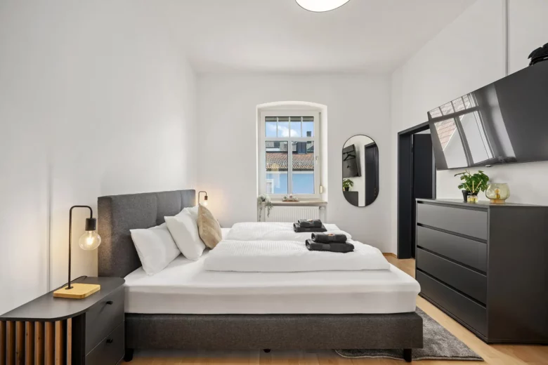 Ein modernes, minimalistisches Schlafzimmer der Ferienwohnung in Bad Kissingen mit einem Doppelbett, Nachttischen und Lampen, Wandspiegel, schwarzer Kommode, TV und einem Fenster für natürliches Licht. Die Handtücher liegen ordentlich auf dem Bett.