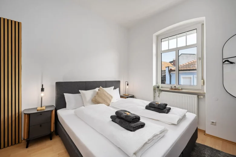 Ein modernes, minimalistisches Schlafzimmer der Ferienwohnung Bachstraße mit einem sauber gemachten Doppelbett, weißen Wänden, zwei Nachttischen mit Lampen, einem großen Fenster, das Tageslicht hereinlässt, und gefalteten Handtüchern auf dem Bett.
