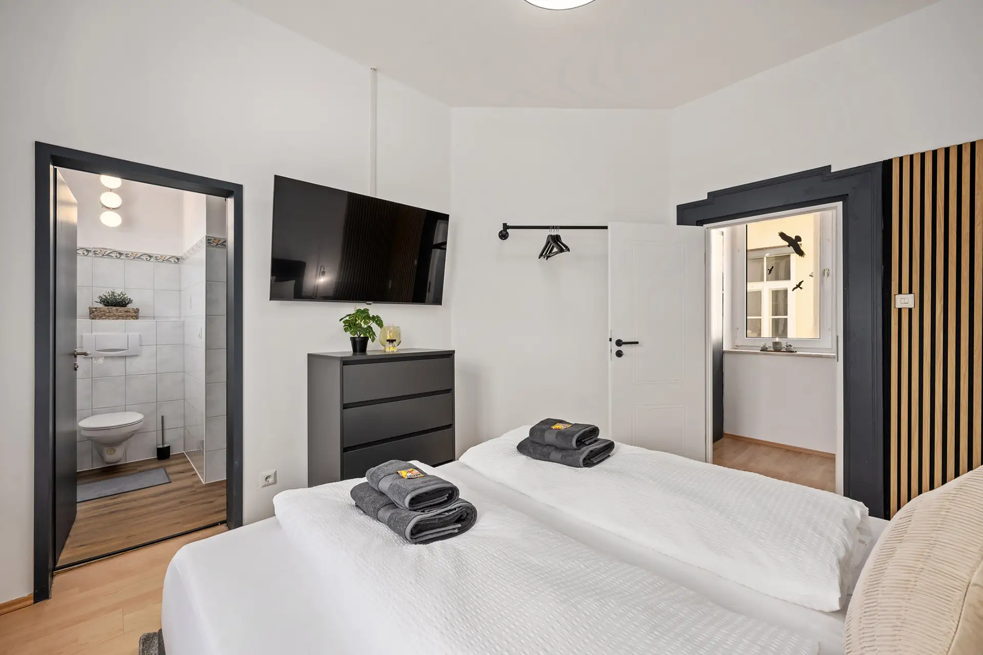 Ein modernes Ferienwohnung-Schlafzimmer in der Bachstraße in Bad Kissingen mit zwei sauber gemachten Einzelbetten, gefalteten Handtüchern, einer schwarzen Kommode mit Pflanzen, einem an der Wand montierten Fernseher und Türen zum Badezimmer und einem kleinen Waschbereich.