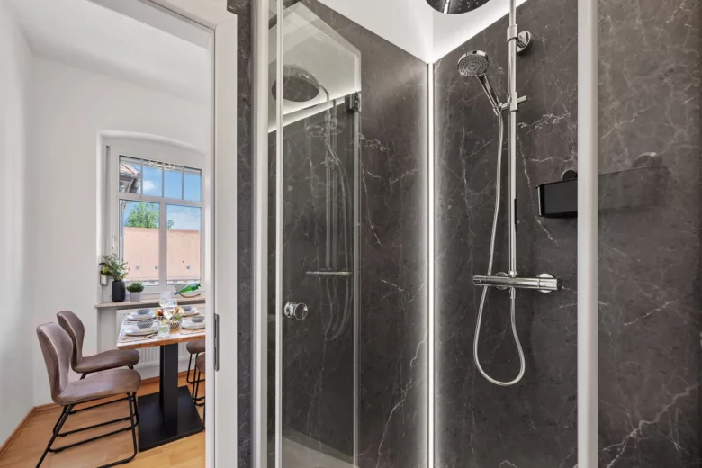 Ein modernes Badezimmer mit einer verglasten Dusche mit Regenbrause und Handbrause. Im Hintergrund ist der Esstisch mit Stühlen der Wohnung durch ein Fenster mit Tageslicht auf der Bachstraße zu sehen.
