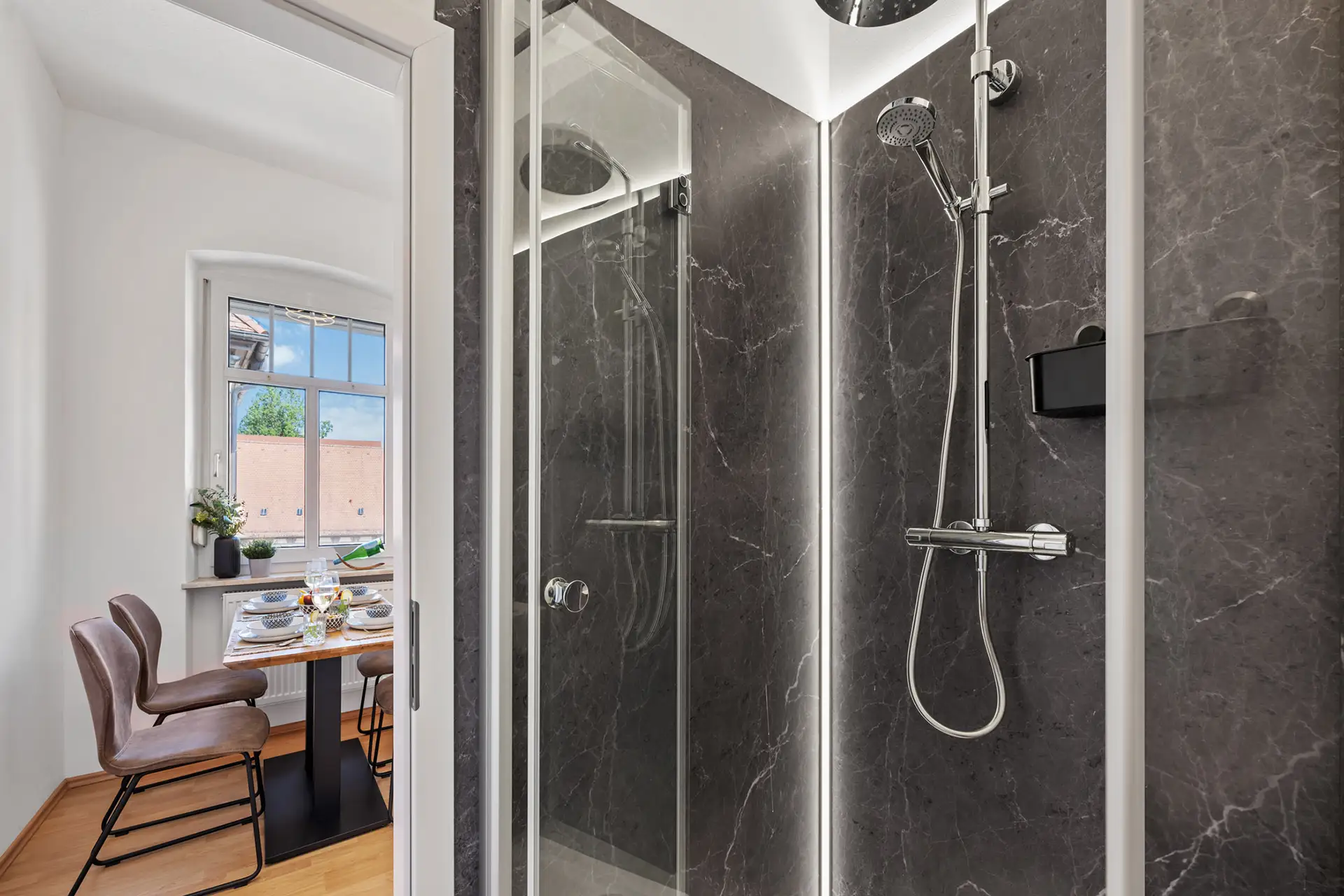 Ein modernes Badezimmer mit einer verglasten Dusche mit Regenbrause und Handbrause. Im Hintergrund ist der Esstisch mit Stühlen der Wohnung durch ein Fenster mit Tageslicht auf der Bachstraße zu sehen.