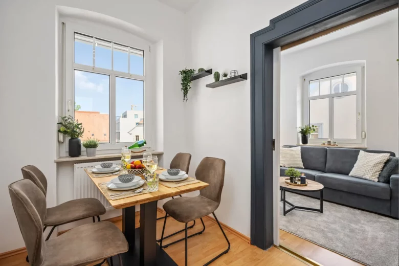 Diese moderne Bad Kissinger Ferienwohnung verfügt über einen Essbereich mit einem Holztisch für vier Personen, braunen Stühlen und einem Fenster mit Topfpflanzen. Durch eine offene Tür gelangt man in ein gemütliches Wohnzimmer mit einem grauen Sofa und einem Couchtisch.