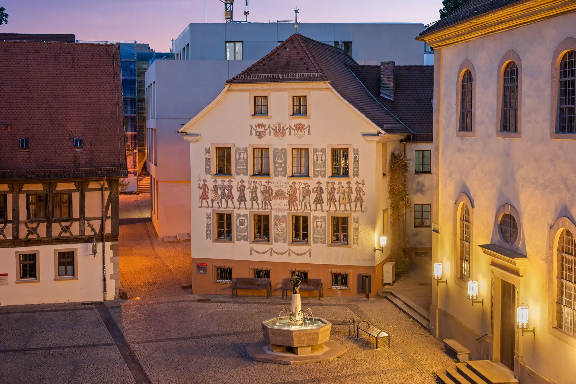 Der kopfsteingepflasterte Rathausplatz in Bad Kissingen in der Abenddämmerung mit einem zentralen Brunnen und historischen Gebäuden - mit gemalten Figuren an der Fassade - die von den Straßenlaternen in der Nähe der charmanten Ferienwohnung sanft beleuchtet werden.