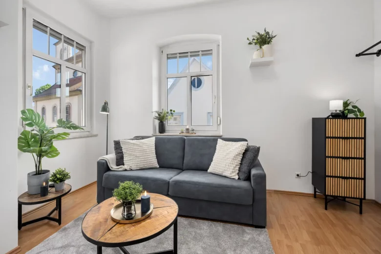 Modernes Wohnzimmer in der Bachstraße mit einem grauen Sofa, zwei hellen Kissen, einem runden Couchtisch aus Holz, Topfpflanzen und großen Fenstern, die natürliches Licht hereinlassen. Helle Holzböden und minimalistische weiße Wände schaffen ein helles, luftiges Gefühl in Bad Kissingen.