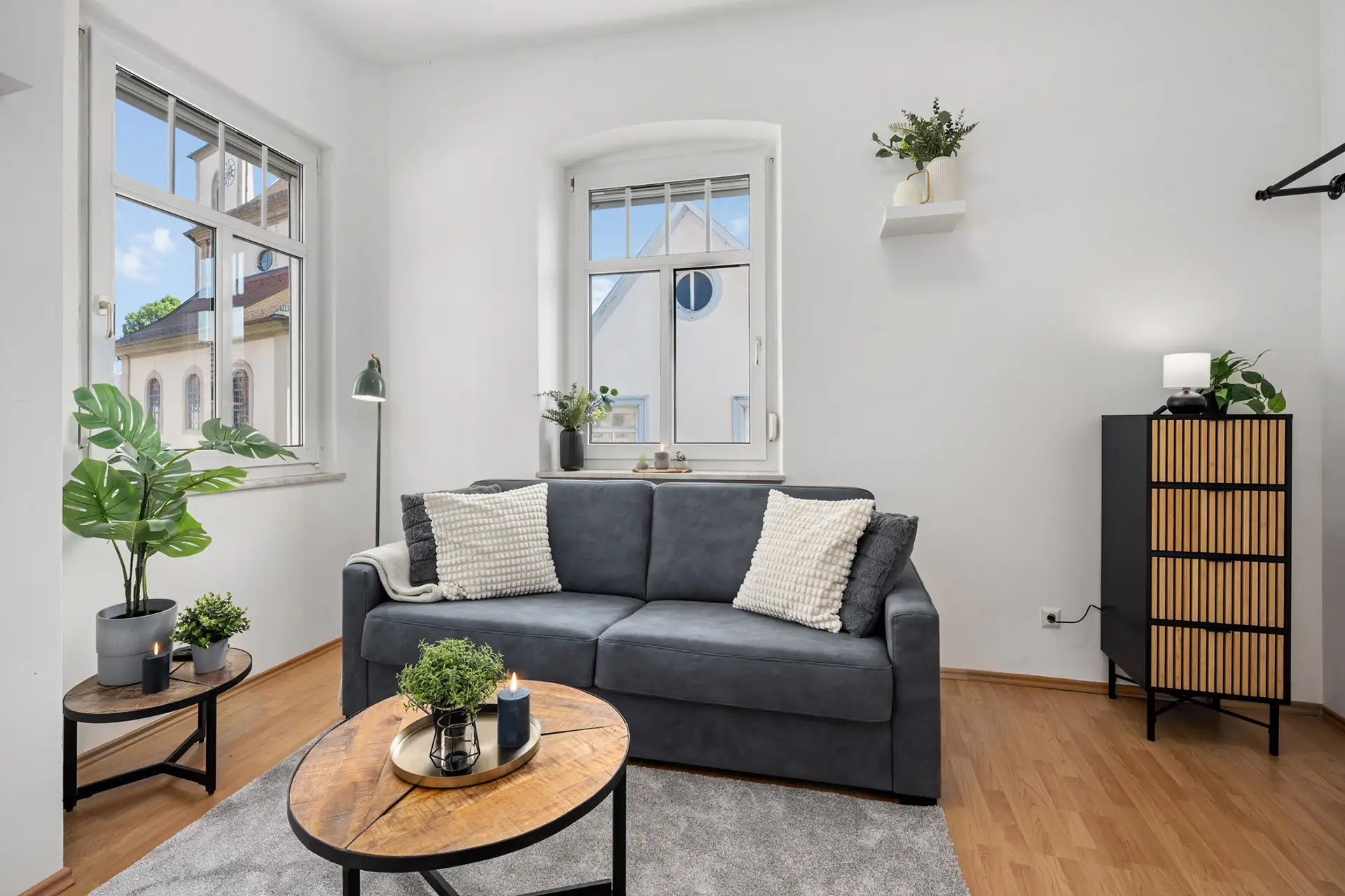 Modernes Wohnzimmer in der Bachstraße mit einem grauen Sofa, zwei hellen Kissen, einem runden Couchtisch aus Holz, Topfpflanzen und großen Fenstern, die natürliches Licht hereinlassen. Helle Holzböden und minimalistische weiße Wände schaffen ein helles, luftiges Gefühl in Bad Kissingen.