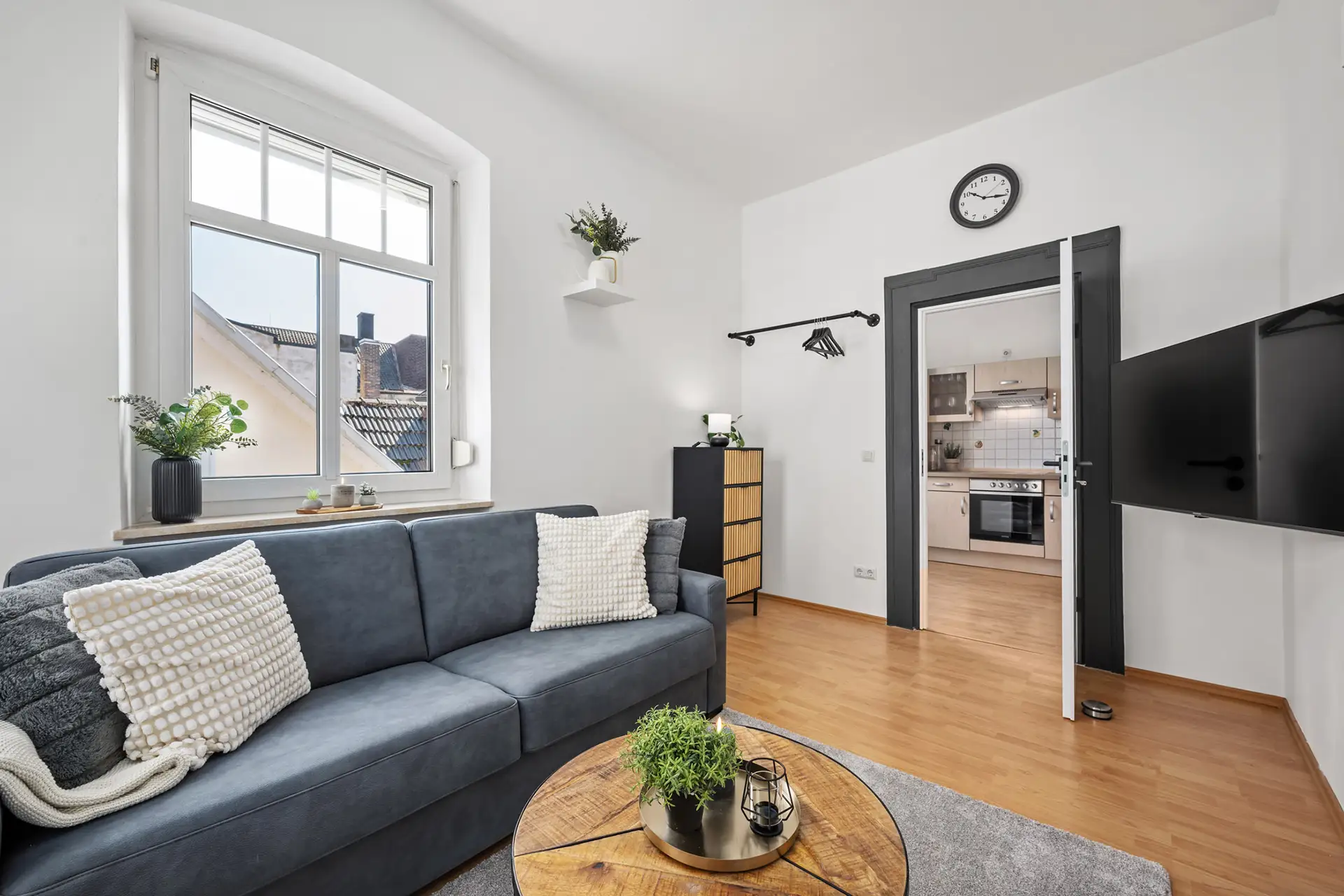 Ein modernes Wohnzimmer mit einem blauen Sofa, weißen Kissen, einem runden Couchtisch aus Holz, Pflanzen und einem an der Wand montierten Fernseher. Eine offene Tür gibt den Blick frei auf eine Küche mit weißen Schränken und Edelstahlgeräten in dieser Ferienwohnung in der Bachstraße in Bad Kissingen.