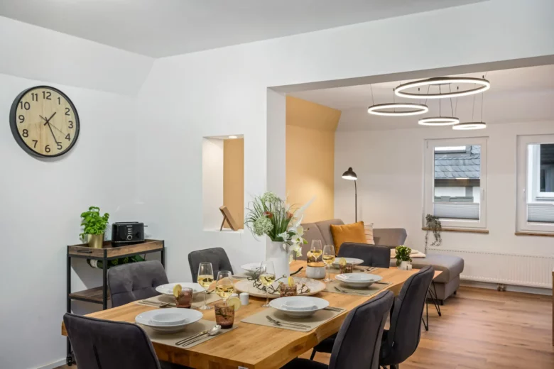 Moderner Ess- und Wohnbereich in der Kirchgasse App.2 mit einem Holztisch für sechs Personen, grauen Stühlen, einem dekorativen Mittelstück und modernen Ringpendelleuchten. Im Hintergrund ein gemütlicher Wohnbereich mit einem grauen Sofa und einer Stehlampe.