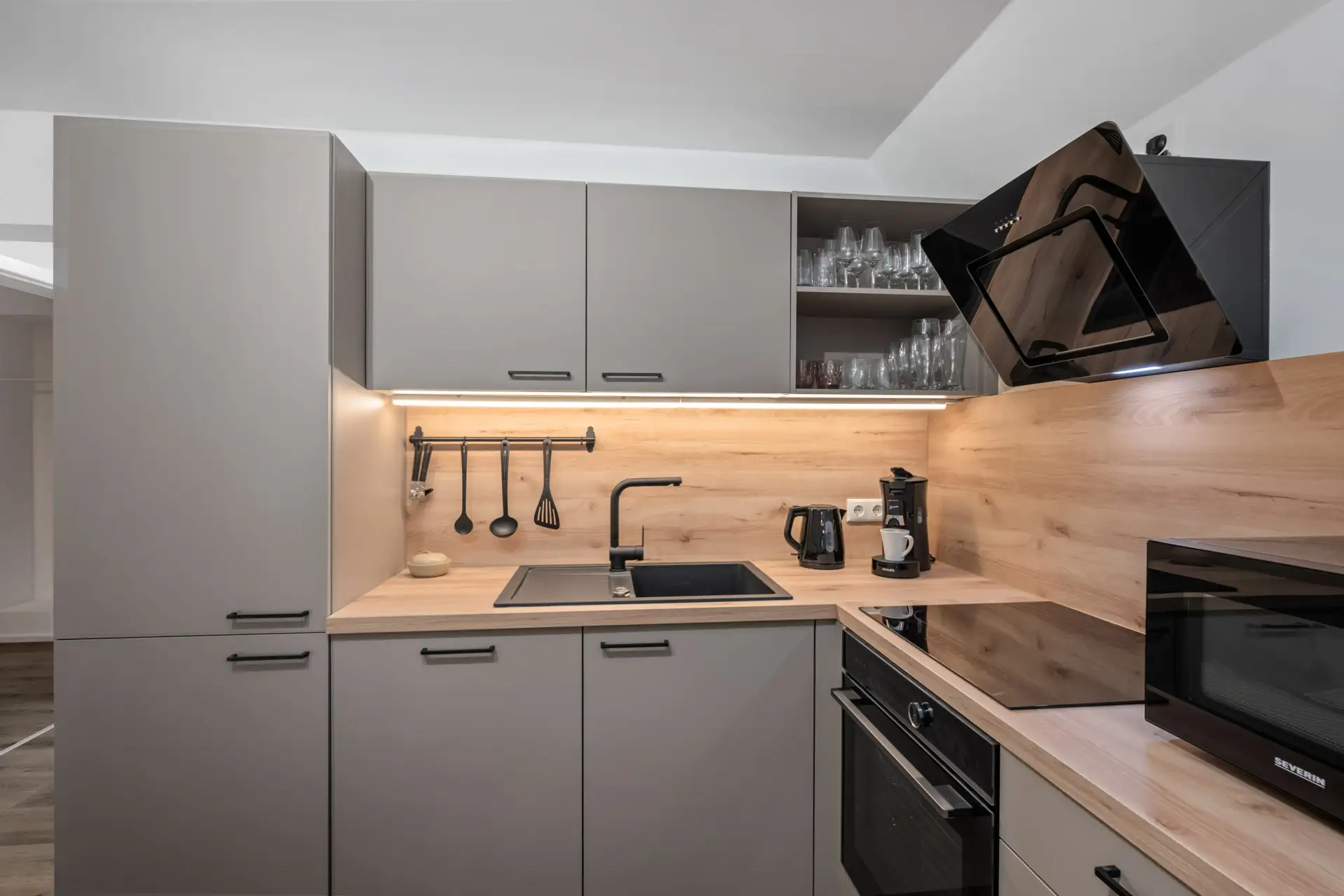 Moderne Küche im App.2 Kirchgasse mit grauen Schränken, schwarzen Geräten, Dunstabzugshaube, Mikrowelle, Backofen, Spüle und Holzarbeitsplatte. Utensilien, Gläser, Wasserkocher und Kaffeemaschine sind übersichtlich angeordnet.