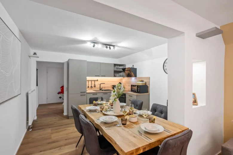 Moderne Essecke und Küche in App.2 Kirchgasse mit einem Holztisch für sechs Personen, grauen Polsterstühlen, neutralen Schränken und heller Beleuchtung. Blumen und Geschirr sind ordentlich auf dem Tisch in dieser stilvollen Wohnung arrangiert.