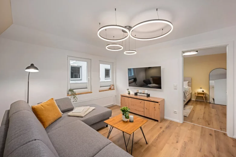 Modernes Wohnzimmer in App.2, Kirchgasse, mit einem grauen Sektionssofa, gelben Akzentkissen, einem Couchtisch aus Holz, einem großen Fernseher auf einer Holzkonsole und runden Deckenleuchten. Das Schlafzimmer ist durch eine offene Tür sichtbar. Helle Holzböden und weiße Wände.