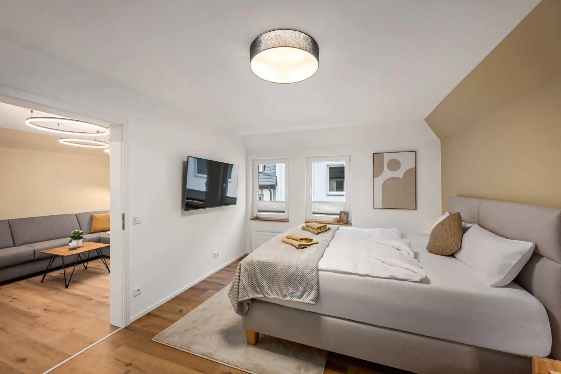 Ein modernes Schlafzimmer in App.2, Kirchgasse verfügt über ein großes Bett mit neutraler Bettwäsche und einer beigen Akzentwand. Zwei Fenster, ein an der Wand montierter Fernseher und minimalistische Kunstwerke ergänzen den Raum. Eine offene Tür führt zu einem Wohnbereich mit einem grauen Sofa und einem Holztisch.