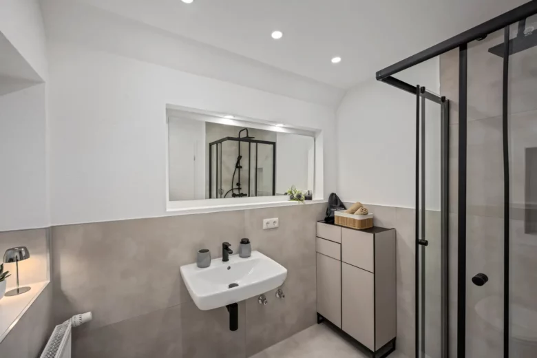 Modernes Badezimmer in App.2, Kirchgasse mit grauen und weißen Tönen, einem wandmontierten Waschbecken mit schwarzen Armaturen, beigem Schrank, Glasduschabtrennung mit schwarzem Rahmen, eingelassener Beleuchtung und einem breiten Innenfenster über dem Waschbecken.