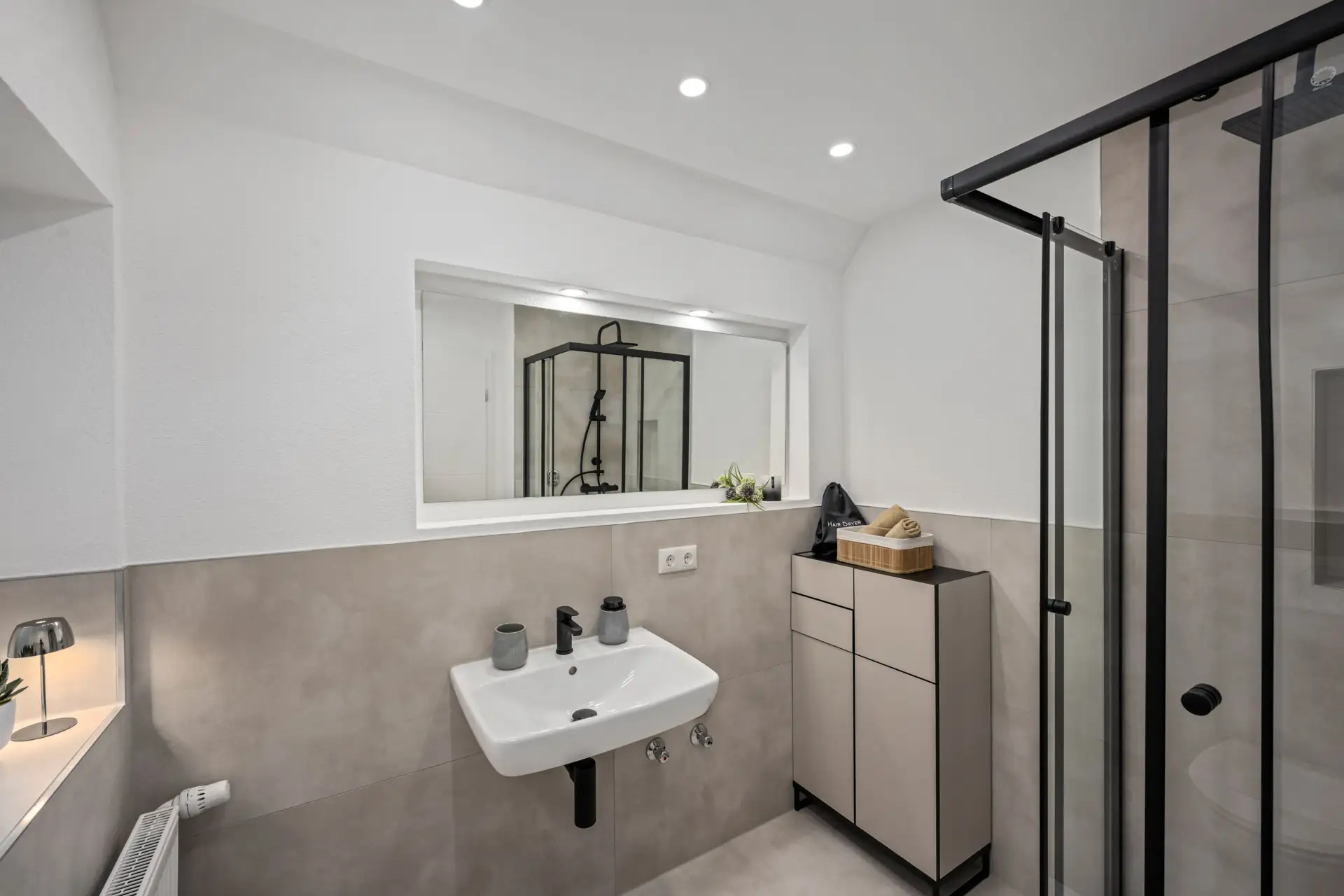 Modernes Badezimmer in App.2, Kirchgasse mit grauen und weißen Tönen, einem wandmontierten Waschbecken mit schwarzen Armaturen, beigem Schrank, Glasduschabtrennung mit schwarzem Rahmen, eingelassener Beleuchtung und einem breiten Innenfenster über dem Waschbecken.