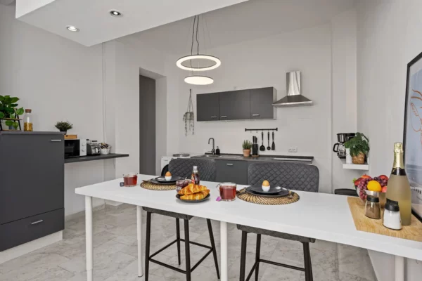 Moderne Küche in der Unteren Marktstraße: graue Schränke, ein weiß gedeckter Tisch für das Frühstück mit Croissants, Saft und Kaffee. Zwei Stühle und ein Tablett mit Sekt und Obst sorgen für Charme - eine einladende Ferienwohnung mit minimalistischem Dekor und sanfter Beleuchtung.