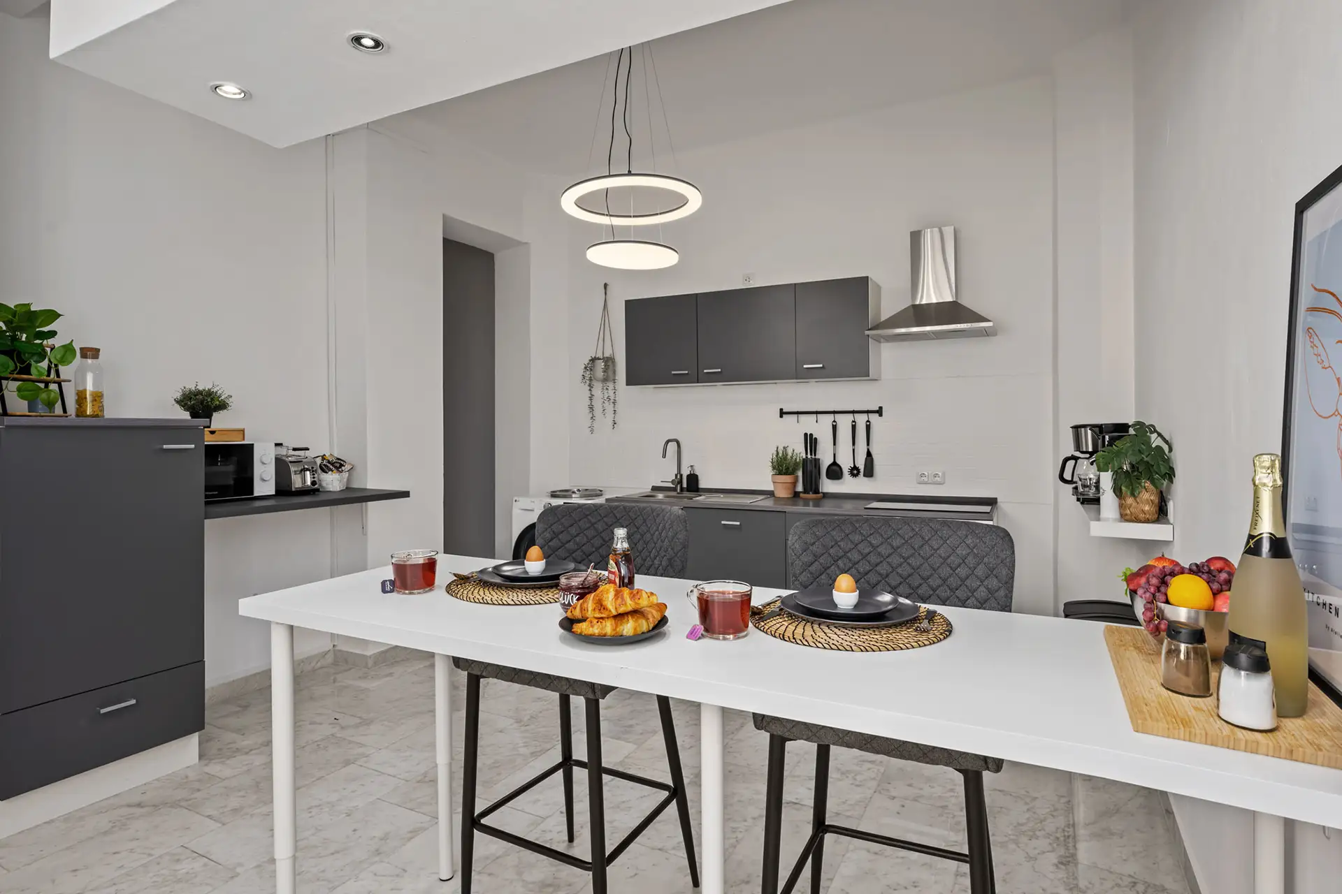 Moderne Küche in der Unteren Marktstraße: graue Schränke, ein weiß gedeckter Tisch für das Frühstück mit Croissants, Saft und Kaffee. Zwei Stühle und ein Tablett mit Sekt und Obst sorgen für Charme - eine einladende Ferienwohnung mit minimalistischem Dekor und sanfter Beleuchtung.
