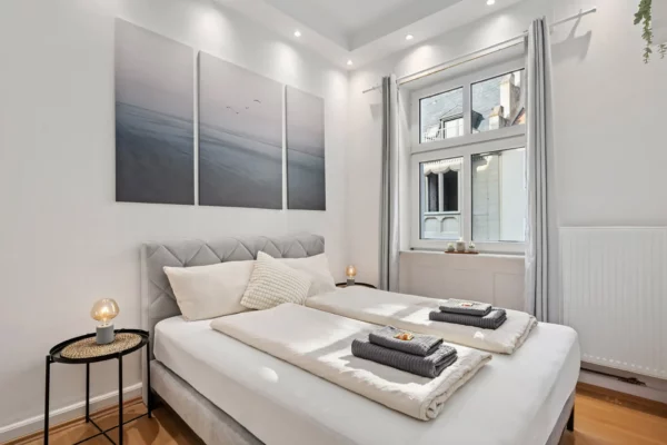 Ein helles, modernes Appartment-Schlafzimmer in der Unteren Marktstraße mit weißen Wänden, einem großen Fenster, einem hellgrauen Polsterbett, das mit weißer Bettwäsche und Handtüchern ausgestattet ist, sowie einer minimalistischen Einrichtung, zu der auch ein dreiteiliges Kunstwerk mit Meeresmotiven über dem Bett gehört.