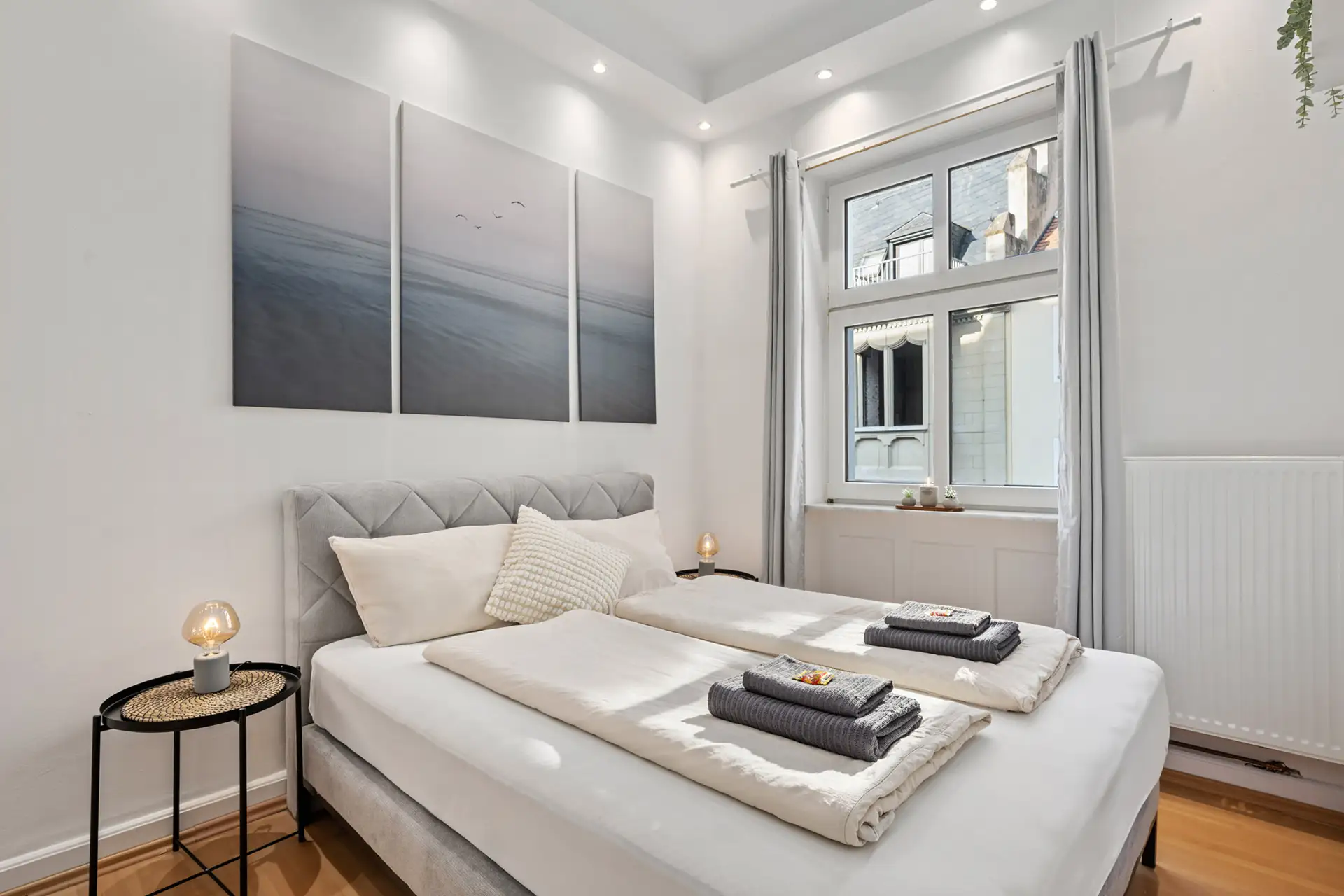 Ein helles, modernes Appartment-Schlafzimmer in der Unteren Marktstraße mit weißen Wänden, einem großen Fenster, einem hellgrauen Polsterbett, das mit weißer Bettwäsche und Handtüchern ausgestattet ist, sowie einer minimalistischen Einrichtung, zu der auch ein dreiteiliges Kunstwerk mit Meeresmotiven über dem Bett gehört.