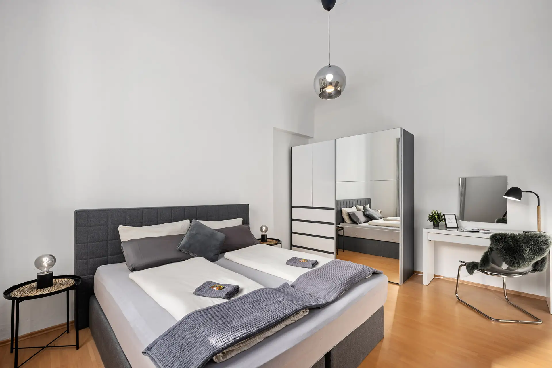 Modernes Appartment-Schlafzimmer in Bad Kissingen in der Unteren Marktstraße mit grauem Polsterbett, weißer und grauer Bettwäsche, verspiegeltem Kleiderschrank, zwei gemütlichen Beistelltischen mit Lampen, einem Schreibtisch mit Stuhl und Lampe und stilvollem Holzfußboden.