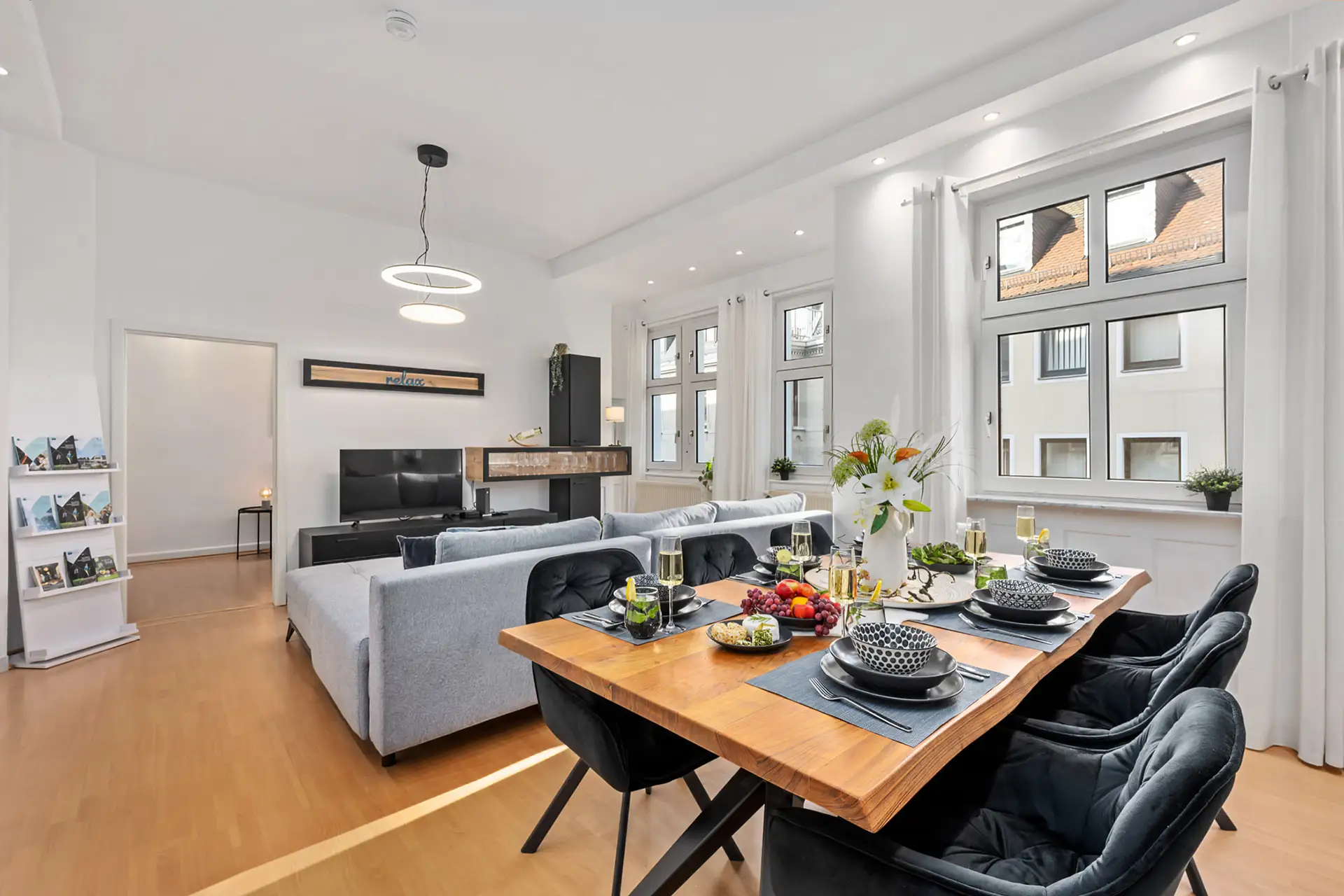 Modernes, offenes Wohn- und Esszimmer in dieser Ferienwohnung in der Unteren Marktstraße, Bad Kissingen, mit hellem Holzfußboden, einem Holztisch für vier Personen, grauen Sofas, großen Fenstern mit weißen Vorhängen und hellem Tageslicht.