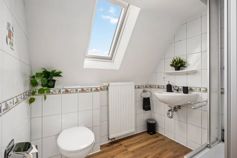 Ein kleines, weiß gefliestes Bad im Appartment in der Bachstraße, Bad Kissingen, mit Dachschräge und Oberlicht, ausgestattet mit Toilette, Waschbecken mit Ablage, Heizkörper, Holzboden und Topfpflanzen zur Dekoration.