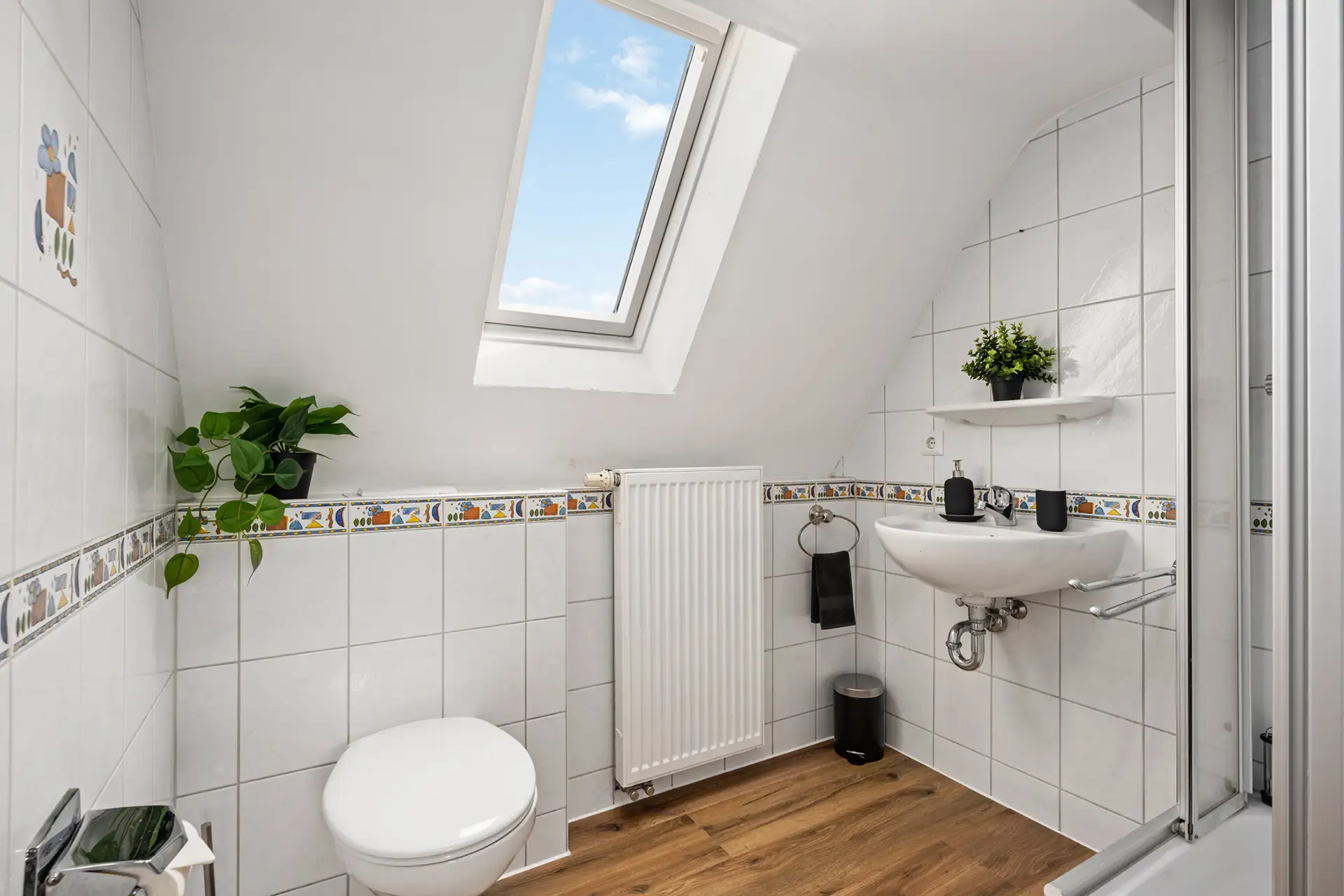 Ein kleines, weiß gefliestes Bad im Appartment in der Bachstraße, Bad Kissingen, mit Dachschräge und Oberlicht, ausgestattet mit Toilette, Waschbecken mit Ablage, Heizkörper, Holzboden und Topfpflanzen zur Dekoration.
